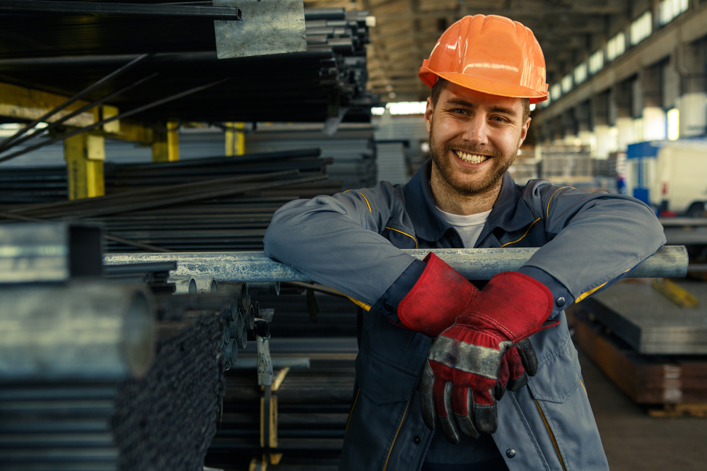 Smiling worker in an orange hard hat and red work gloves leaning on a steel pipe in a warehouse, representing confident smiles in Decatur, Alabama